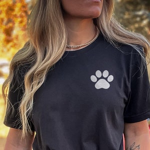 May include: A black t-shirt with a white paw print design. The shirt is being worn by a person with long blonde hair. The person is sitting on a red ATV.