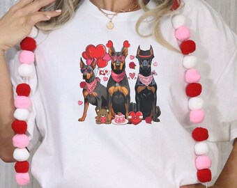 May include: White t-shirt featuring a graphic of three Doberman Pinschers wearing bandanas and sunglasses, surrounded by hearts and a teddy bear. The shirt is paired with pink shorts and a red, pink, and white pom-pom garland.