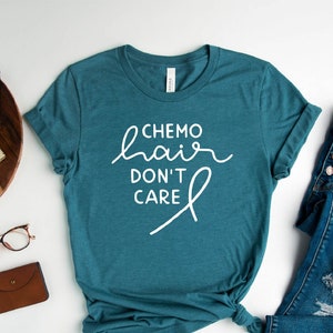 May include: A teal blue t-shirt with the text "Chemo hair don't care" in white lettering and a white ribbon design. The shirt is laid flat on a white surface with a pair of glasses, a brown wallet, a wooden tray with a camera and watch, and a pair of blue jeans and white trainers.
