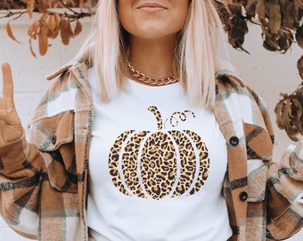 Puede incluir: Una camiseta blanca con un gráfico de calabaza de estampado de leopardo. La modelo lleva leggings negros y botas marrones. También lleva una camisa de cuadros marrones y blancos y una boina marrón.