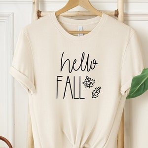 May include: A beige t-shirt with the text "hello fall" and a small leaf design.