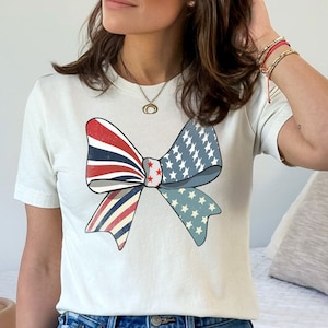 May include: White t-shirt with a red, white, and blue bow graphic. The bow is made of an American flag pattern.