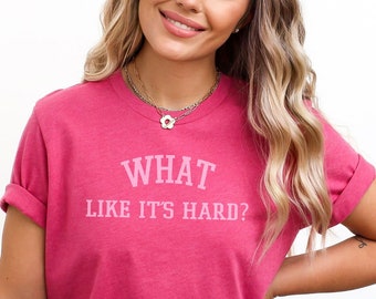 Peut inclure: Un t-shirt rose avec le texte "WHAT LIKE IT'S HARD?" en lettres blanches.