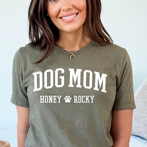 May include: An olive green t-shirt featuring the white text "DOG MOM", with "HONEY ROCKY" and a paw print underneath. The t-shirt is worn with blue denim jeans.