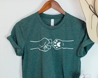 May include: A dark green t-shirt with a white graphic of a fist bumping a paw print. The t-shirt is hanging on a wooden hanger against a white wall.