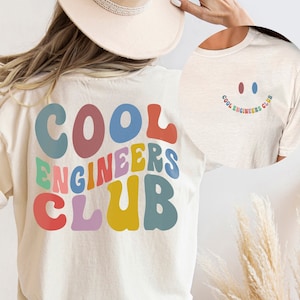 May include: A white t-shirt with the text "COOL ENGINEERS CLUB" in rainbow colored letters. The letters are in a retro font style. The shirt is being worn by a person with long blonde hair and a white hat.