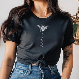 May include: A black t-shirt with a white bee graphic and the word "lett" written vertically below it. The shirt is being worn by a person with a tattoo on their arm.