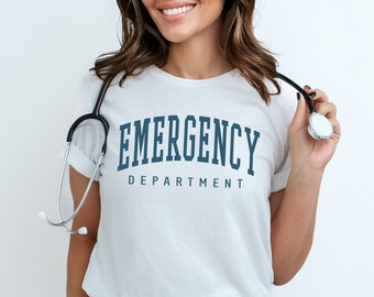 Op de afbeelding: Een wit T-shirt met de tekst "EMERGENCY DEPARTMENT" in blauwe letters. De persoon draagt een zwarte broek en een stethoscoop om de nek.
