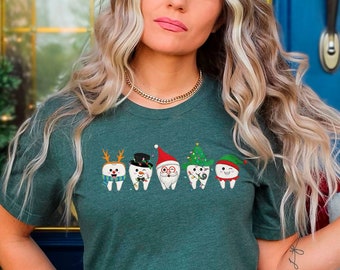 May include: A green t-shirt with a festive design of five teeth wearing Christmas hats and accessories. The teeth are wearing a reindeer hat, a top hat, a Santa hat, a Christmas tree hat, and an elf hat.