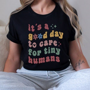 May include: A black t-shirt with the text "it's a good day to care for tiny humans" in colorful retro-style lettering.