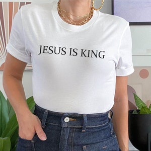 Pode incluir: Camiseta branca com o texto "JESUS IS KING" impresso em preto. A camiseta est&aacute; sendo usada por uma pessoa que est&aacute; usando shorts jeans azuis.