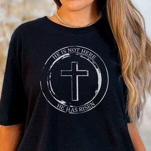 K&ouml;nnte beinhalten: Schwarzes T-Shirt mit einem wei&szlig;en Kreis-Motiv, das ein Kreuz und den Text "Er ist nicht hier, Er ist auferstanden" zeigt.