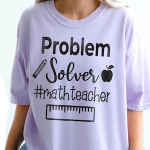 Può includere: T-shirt lilla chiaro con le scritte nere "Problem Solver" e "#mathteacher". Il design include una matita, una mela e un righello. La maglietta è in stile casual.