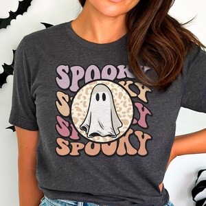 Puede incluir: Una camiseta gris oscuro con un gr&aacute;fico de un fantasma blanco en un c&iacute;rculo rosa, morado y marr&oacute;n con la palabra "SPOOKY" repetida alrededor del c&iacute;rculo.