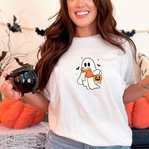 May include: White t-shirt with a graphic of a ghost holding a book and a pumpkin. The ghost is wearing a witch hat. The shirt is on a person wearing blue jeans.