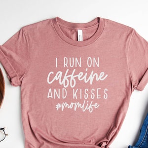 K&ouml;nnte beinhalten: Ein T-Shirt in Heide-Mauve mit dem Aufdruck "I run on caffeine and kisses #momlife" in Wei&szlig;.