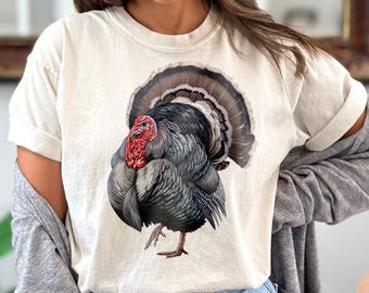 Puede incluir: Camiseta color marfil con una ilustración detallada de un pavo. El pavo tiene plumas negras, grises y marrones, una carúncula roja y una cola en forma de abanico. La camiseta es de corte holgado y tiene las mangas enrolladas. La palabra "IVORY" está en un círculo en la esquina superior derecha.