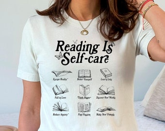 May include: White t-shirt with the text "Reading Is Self-care" and illustrations of books with different phrases like "Escape Reality", "Better Yourself", "Learn New Things", "Fall in Love", "Think Deeper", "Discover New Worlds", "Admire Auctry", "Find Passion", "Make New Friends".