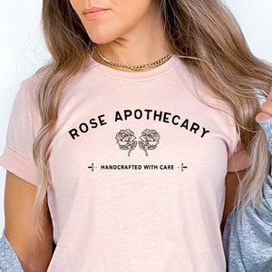 May include: A light pink t-shirt with the text "ROSE APOTHECARY" in black, arched above two rose illustrations. Below the roses is the text "HANDCRAFTED WITH CARE". The model is wearing blue denim shorts and a gray cardigan.