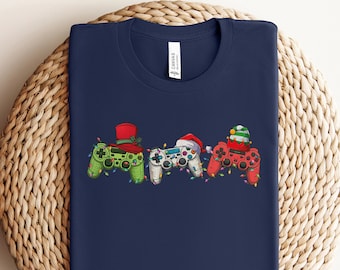 Könnte beinhalten: Marineblaues T-Shirt mit einem Motiv von drei Videospielcontrollern, die Weihnachtsmützen tragen. Die Controller sind grün, weiß und rot.