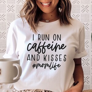 K&ouml;nnte beinhalten: Wei&szlig;es T-Shirt mit schwarzem Text, der "I run on caffeine and kisses #momlife" lautet. Eine Person tr&auml;gt das Shirt und h&auml;lt eine wei&szlig;e Tasse.