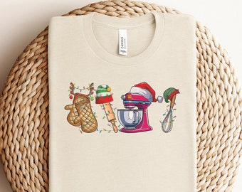 May include: A beige t-shirt with a Christmas-themed design featuring a red stand mixer, a rolling pin, a red and green oven mitt, and a green whisk. The design is surrounded by Christmas lights.