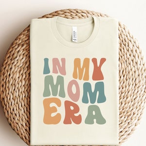 May include: A beige t-shirt with the text "In My Mom Era" in colorful, retro-style lettering.