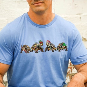 May include: Light blue t-shirt with a festive design featuring four turtles adorned with Christmas lights, scarves, and Santa hats. The shirt has the text "Heather Columbia" on the upper left and the brand logo "Oje Apparel" on the upper right.