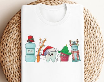 May include: White t-shirt with a Christmas-themed dental hygiene design. The design features a tooth wearing a Santa hat, a toothbrush with lights, a bottle of mouthwash, a tube of toothpaste with a reindeer, and a small Christmas tree.
