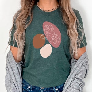 Puede incluir: Una camiseta verde oscuro con un dise&ntilde;o gr&aacute;fico de formas abstractas en rosa, marr&oacute;n y blanco. El dise&ntilde;o presenta una hoja estilizada.
