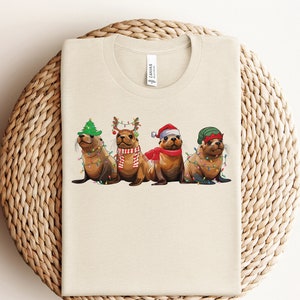 May include: A beige t-shirt with a Christmas-themed design featuring three sea lions wearing festive hats and lights. The sea lions are wearing a Santa hat, a reindeer headband, and a green elf hat.