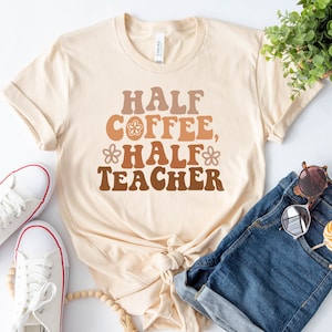 May include: A beige t-shirt with the text "Half Coffee, Half Teacher" in brown, orange, and yellow lettering with a flower design. The shirt is laid flat on a white surface with a pair of white sneakers, a wooden bead necklace, a pair of sunglasses, and denim shorts.