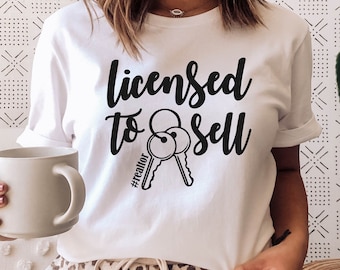Może przedstawiać: Biała koszulka z czarnym napisem "Licensed to Sell #realtor" i grafiką dwóch kluczy. Osoba nosząca koszulkę siedzi na kanapie w panterkę.