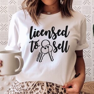 Pode incluir: Camiseta branca com texto preto que diz "Licensed to Sell #realtor" e um gr&aacute;fico de duas chaves. A pessoa que est&aacute; usando a camiseta est&aacute; sentada em um sof&aacute; com estampa de leopardo.
