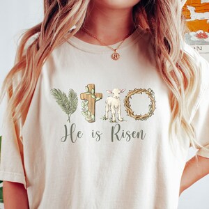 May include: A cream colored t-shirt with a graphic of a cross, a lamb, a palm branch, and a crown of thorns. The text "He is Risen" is printed below the graphic.
