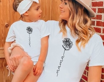 Könnte beinhalten: Ein weißes T-Shirt mit einer schwarzen Sonnenblume und dem Wort "mama" in Kursivschrift. Ein kleineres weißes T-Shirt mit einer schwarzen Sonnenblume und dem Wort "mini" in Kursivschrift. Die Shirts werden von einer Frau und einem jungen Mädchen getragen.