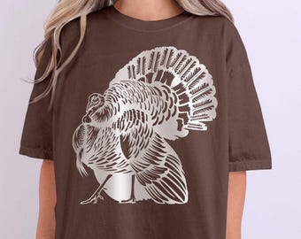 Puede incluir: Camiseta color espresso con un gráfico detallado de un pavo plateado. El diseño muestra las plumas y el cuerpo del pavo en una ilustración estilizada. La camiseta tiene un corte holgado y mangas cortas. La palabra "ESPRESSO" está en un círculo en la esquina superior derecha.