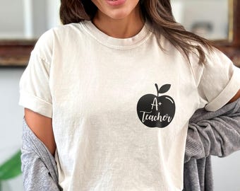 May include: Ivory-coloured t-shirt with a black apple graphic and the text "A+ Teacher." The shirt has short sleeves and a relaxed fit. The model is wearing blue jeans and a grey cardigan.
