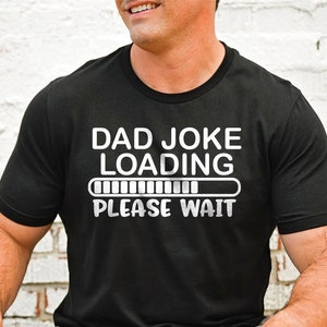 Puede incluir: Camiseta negra con el texto blanco "DAD JOKE LOADING PLEASE WAIT" y una barra de carga. La camiseta es de cuello redondo.