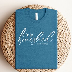 May include: A teal blue t-shirt with the text "It is finished" in white cursive lettering and the scripture reference "John 19:28-30" underneath.
