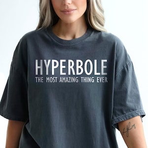May include: Dark grey t-shirt with the word "HYPERBOLE" in large, white, capital letters, and the phrase "THE MOST AMAZING THING EVER" below it. The shirt is a relaxed fit and has short sleeves.