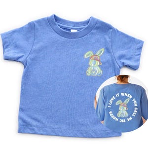 Puede incluir: Una camiseta de manga corta azul claro con un conejito de dibujos animados con una gorra de b&eacute;isbol al rev&eacute;s en la parte delantera. La parte posterior de la camiseta tiene un c&iacute;rculo blanco con el texto "I LOVE IT WHEN YOU CALL ME BIG HOPPA" y un conejito de dibujos animados con una gorra de b&eacute;isbol al rev&eacute;s.