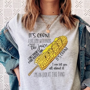 Op de afbeelding: Een grijs T-shirt met een afbeelding van een maïskolf en de tekst "It's Corn! A big lump with knobs & the juice. I can't imagine a more beautiful thing. I mean look at this thing!"
