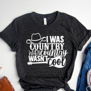 May include: A dark gray t-shirt with a white graphic that says "I was country when country wasn't cool". The graphic includes a cowboy hat and a guitar.
