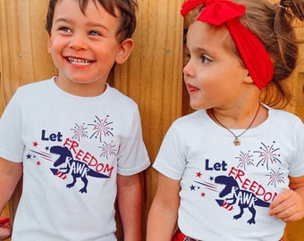 Puede incluir: Dos niños con camisetas blancas con un gráfico de dinosaurio y el texto "Let Freedom Rawk" en rojo, blanco y azul. La niña lleva una diadema roja y una camisa de cuadros rojos y negros atada a la cintura. El niño lleva pantalones vaqueros azules.