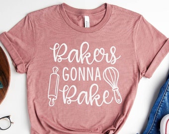 Könnte beinhalten: Ein pinkfarbenes T-Shirt mit dem Text "Bakers Gonna Bake" in weißen Buchstaben. Das Shirt zeigt ein Nudelholz und einen Schneebesen.