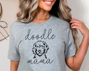May include: A light gray t-shirt with the words "doodle mama" in a script font, with a doodle dog illustration. The shirt is short-sleeved and made of a soft material.