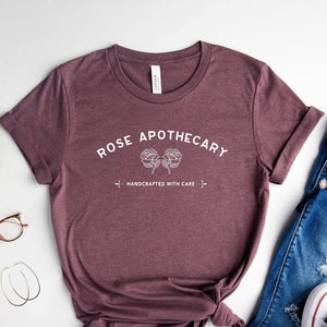 May include: A maroon t-shirt with the text "ROSE APOTHECARY HANDCRAFTED WITH CARE" and a floral design, paired with blue jeans and white sneakers. A wooden tray with a small camera, watch, and glasses is also visible.