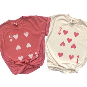 May include: Two t-shirts, one coral and one cream, each with a playing card design. The coral shirt features a six of hearts, and the cream shirt displays a seven of hearts. Both shirts are knotted at the waist.