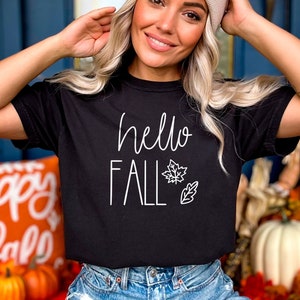 May include: A black t-shirt with the text "hello fall" in white lettering and a white outline of a leaf. The shirt is being worn by a person with blonde hair and a beige beanie.